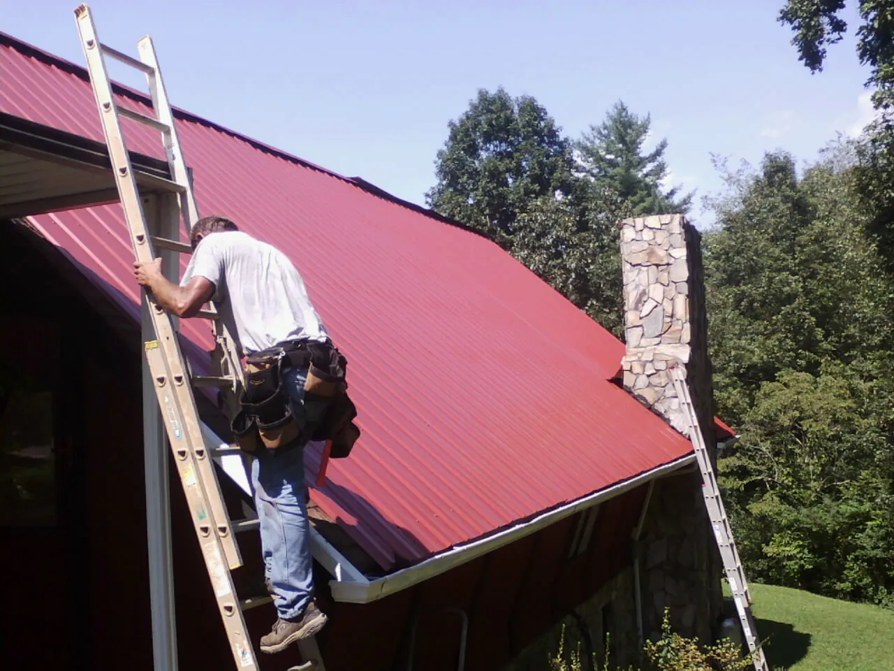 Roofer on a ladder beside a red metal roof and stone chimney — Soffit & Fascia Repair by Roofing Brothers in Hereford